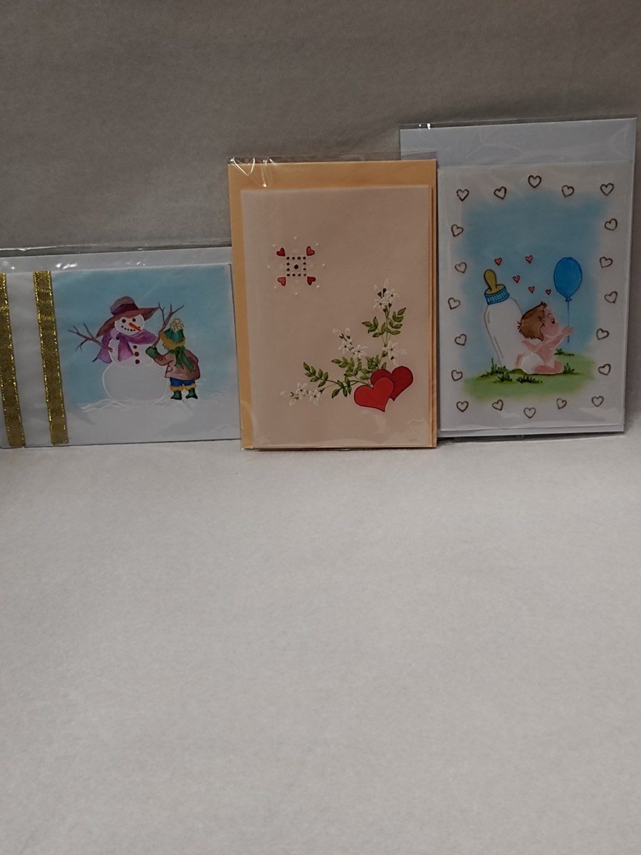 Trois cartes faites main présentées sous pochette transparente avec leur enveloppe : une carte de Noël bleue à bonhomme de neige bordée d’un galon doré, une carte crème à cœurs rouges et petites fleurs blanches, et un faire-part de naissance encadré de cœurs, avec un bébé et un ballon bleu.