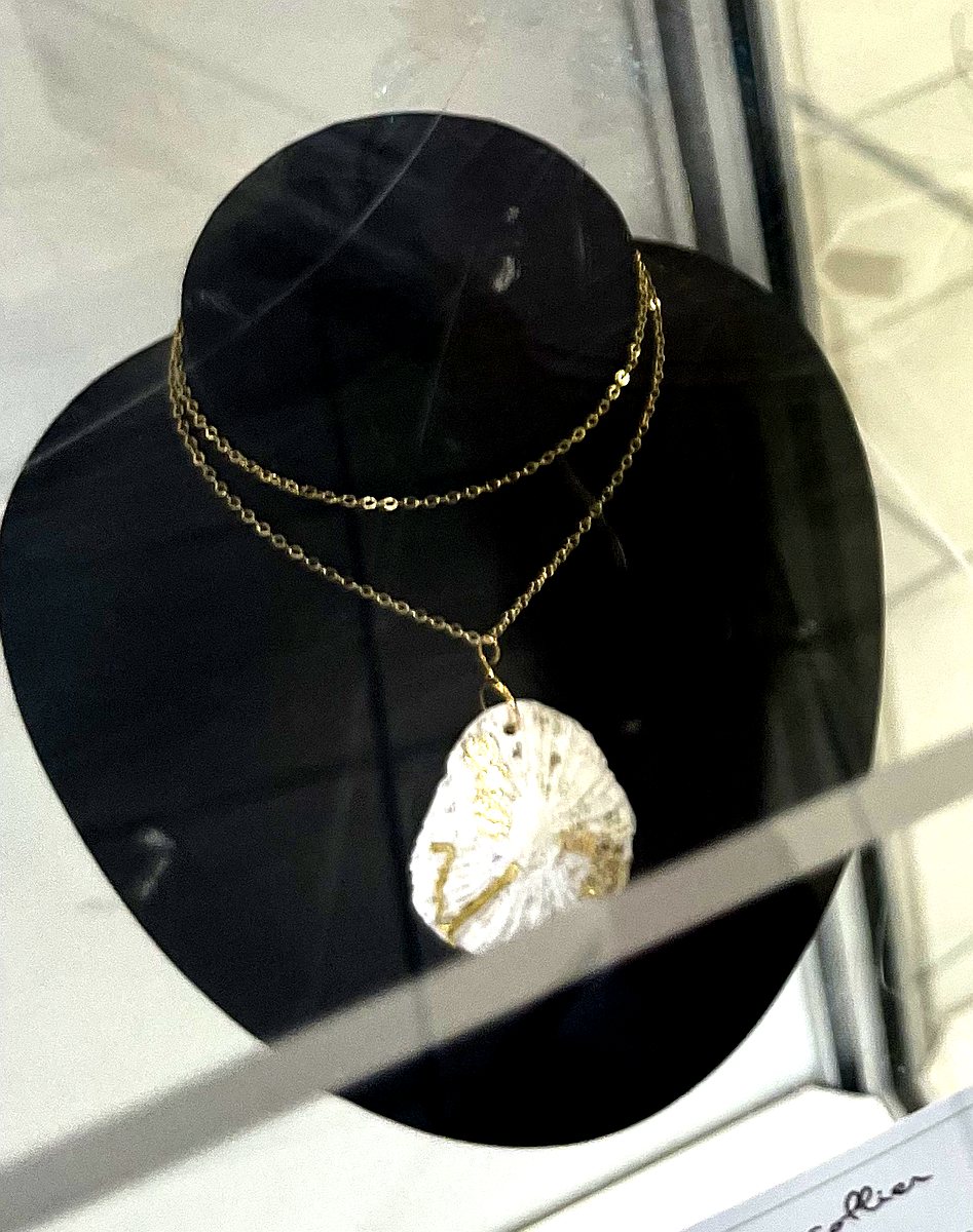 Collier à chaîne dorée double et pendentif coquillage blanc nacré rehaussé d’un liseré doré, présenté sur un buste noir.