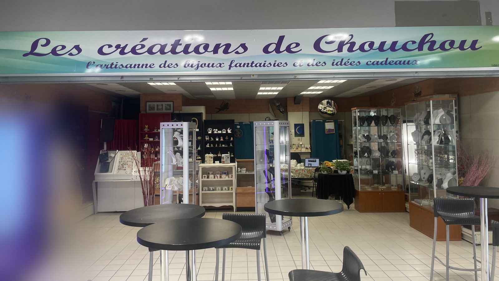 La boutique vue depuis la galerie : l’enseigne « Les créations de Chouchou », suivie de sa baseline manuscrite « l’artisanne des bijoux fantaisies et des idées cadeaux », en lettres cursives violettes sur un bandeau dégradé turquoise et blanc ; en dessous, les vitrines éclairées et les présentoirs de bijoux.