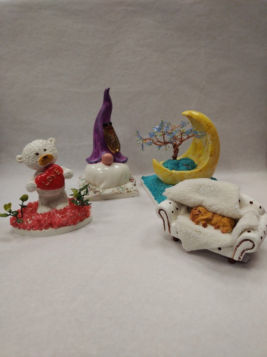 Quatre figurines de décoration en plâtre faites main : un ourson blanc serrant un cœur rouge marqué LOVE sur un socle rouge pailleté, un gnome au long bonnet violet, une lune jaune portant un petit arbre fleuri sur un lit de granulés turquoise, et un chien couché sur un canapé blanc.