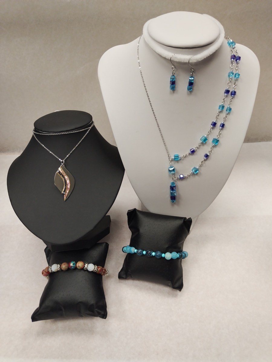 Quatre bijoux faits main présentés ensemble : un collier deux rangs de perles de verre carrées bleu turquoise et bleu nuit sur chaîne argentée avec ses boucles d’oreilles assorties, un pendentif en forme de feuille argentée à reflets nacrés, et deux bracelets de perles, l’un en pierres beige et brique, l’autre en perles turquoise.