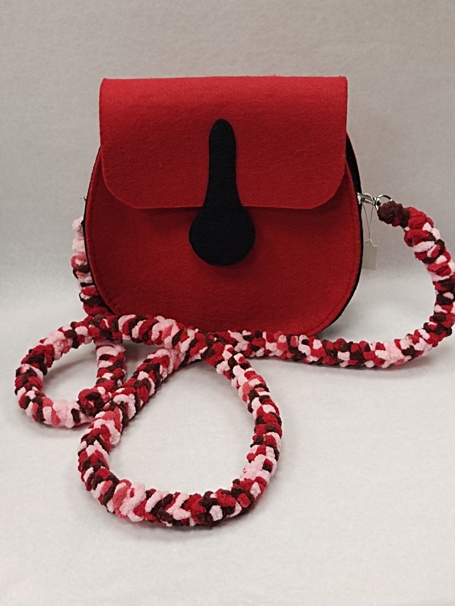 Sac à main en feutrine rouge fait main, rabat arrondi orné d’un motif noir en forme de goutte, porté par une longue anse épaisse tressée à la main en laine rouge, rose et blanche.